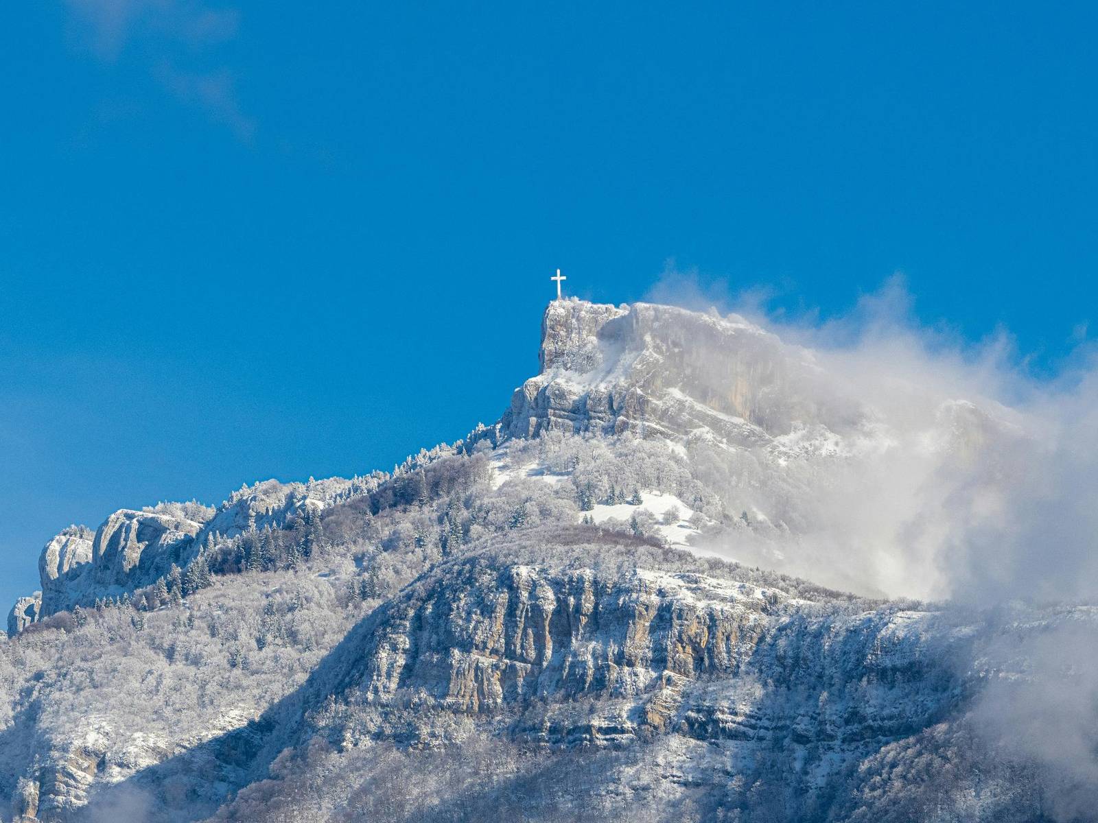La croix du Nivolet au sommet d’une falaise enneigée du massif des Bauges, sous un ciel bleu, au-dessus de Chambéry